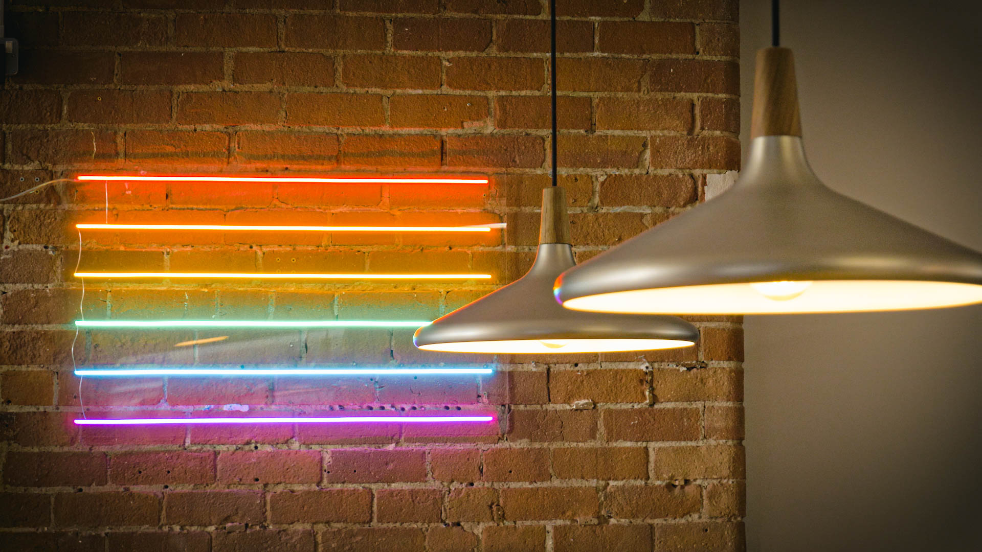 Photograph of a neon rainbow sign on the wall of the EMEA Recruitment office kitchen, with two ceiling lights hanging down in front of it