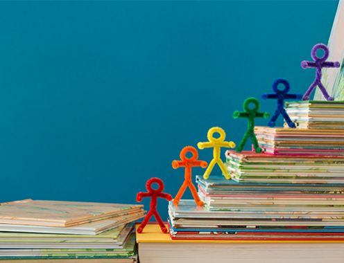 Photograph of a stack of books creating steps, with multi-colored stick people made from pipe cleaners on each step
